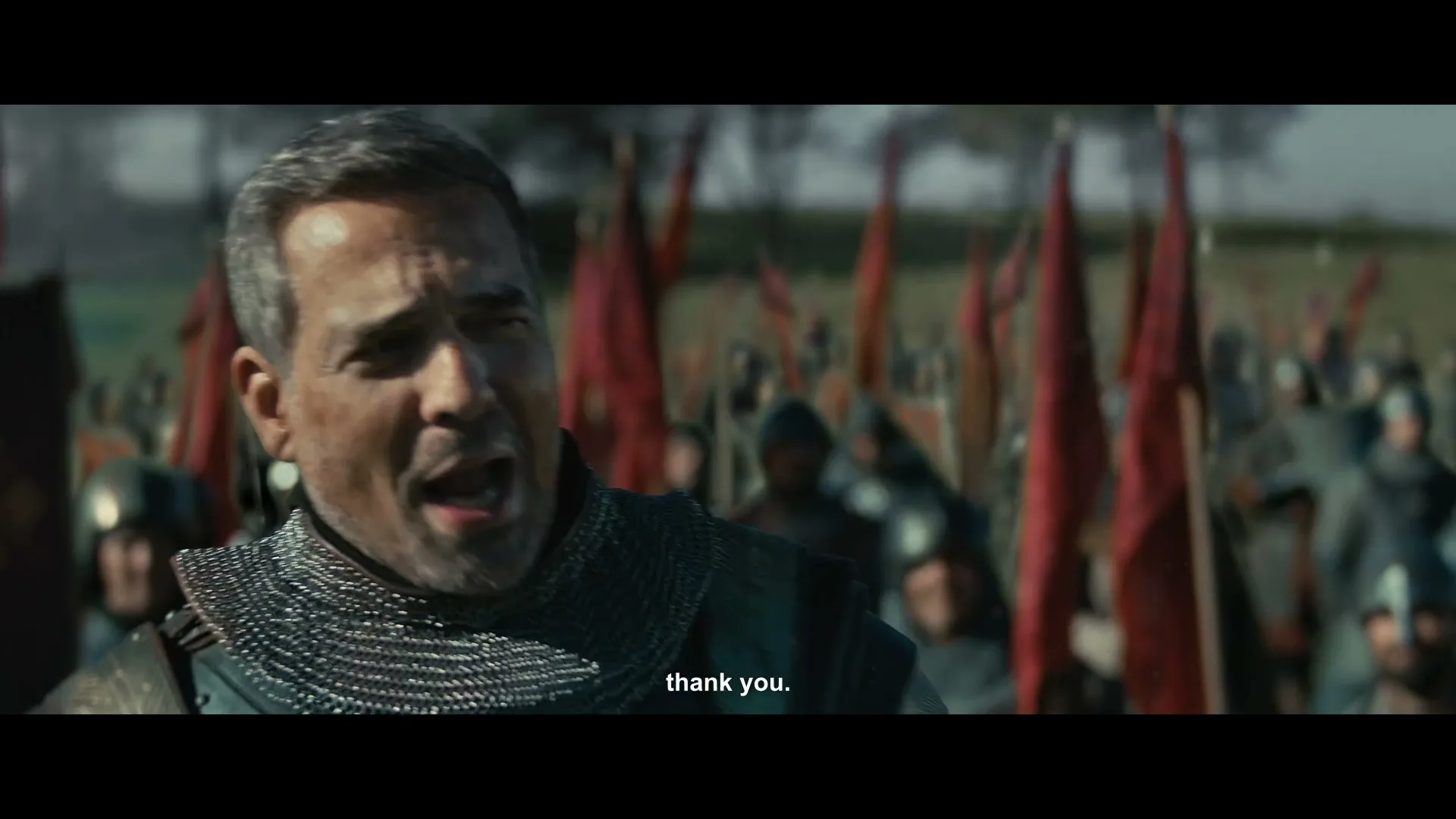 A medieval commander in chainmail armor, with a gray beard, shouts and smiles at his troops on a battlefield.