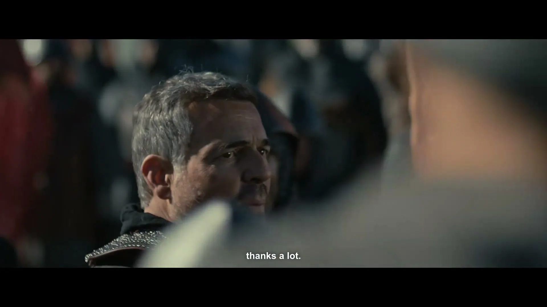 A middle-aged man with grey hair and a beard, wearing medieval armor, looks around a battlefield filled with other armored soldiers.