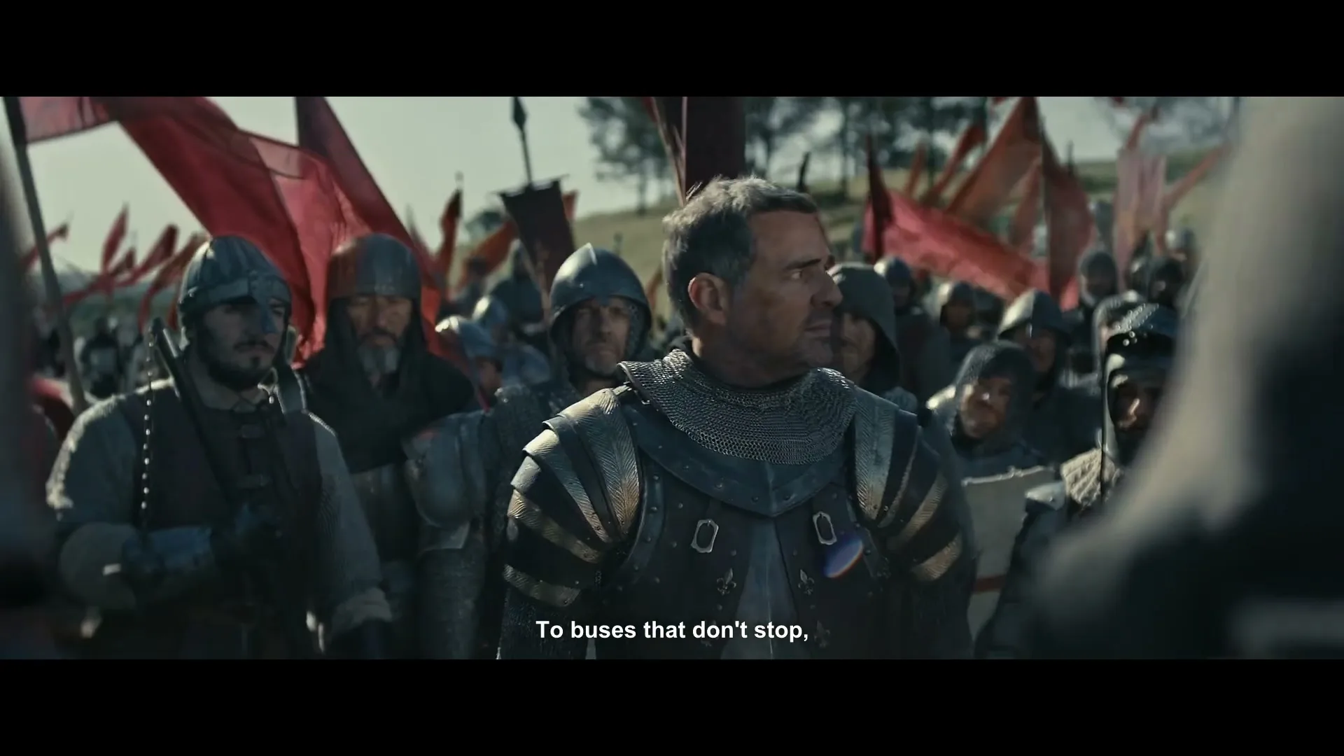 A male knight in armor, surrounded by other knights and red flags, looks around with a determined expression on a battlefield.