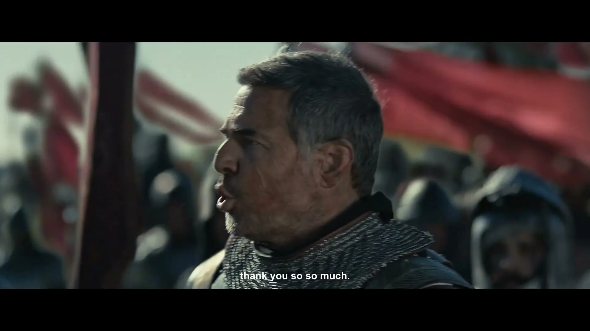 A battle-worn male warrior in chainmail armor, with scars on his face, looks around a battlefield with red flags in the background.