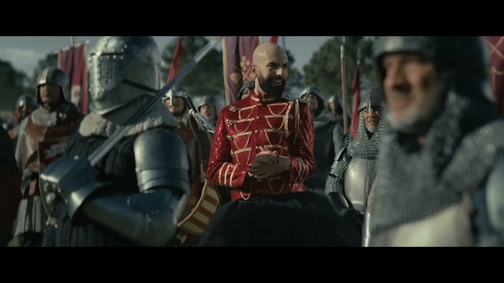 A bald man with a beard in a sparkly red military jacket stands among medieval knights on a battlefield, looking around with a slight smile.
