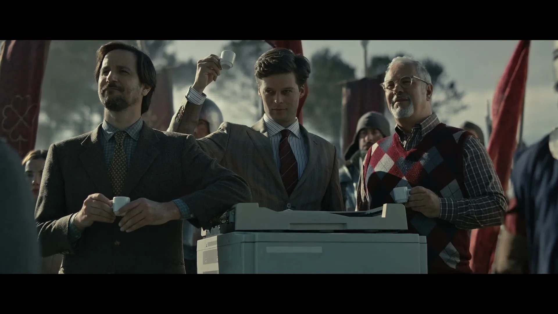 Three men in business attire stand around a printer, holding small coffee cups, surrounded by people in medieval armor on a battlefield.