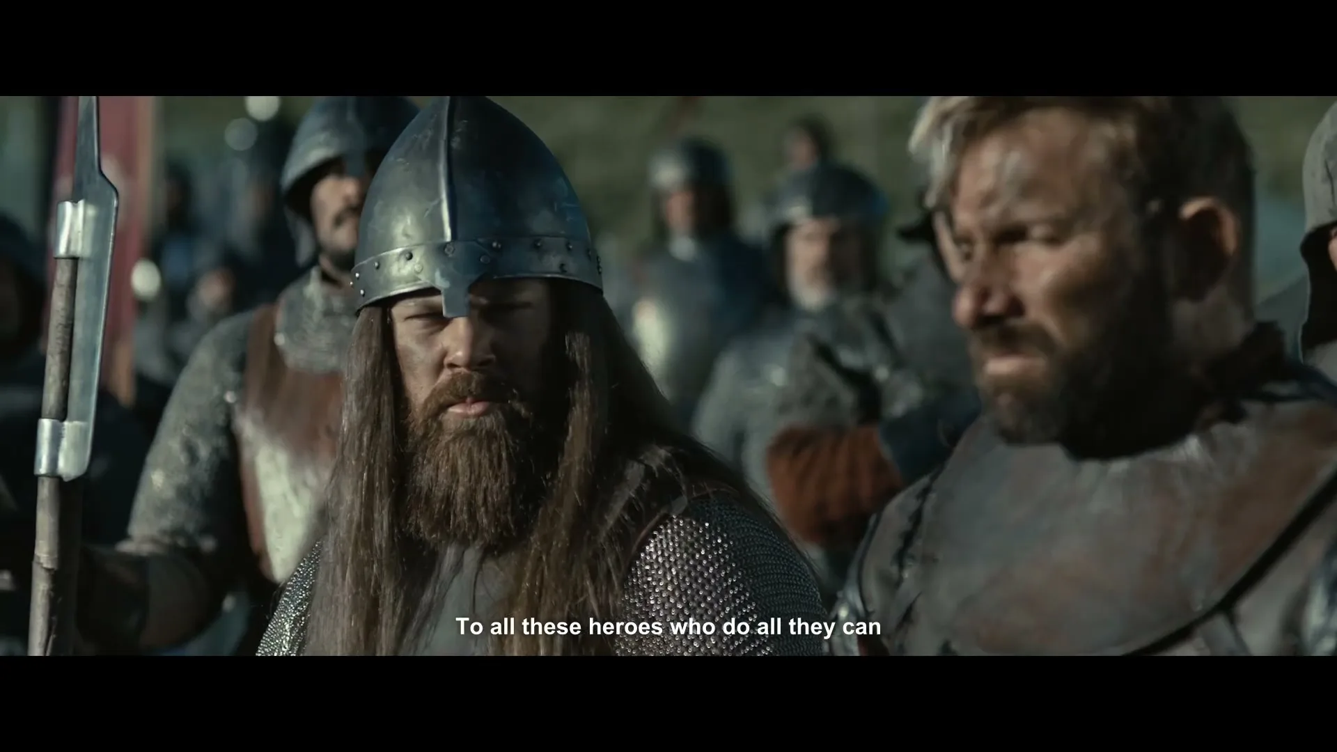 Two male warriors, one with a helmet and long hair, the other with a beard and short hair, stand among other warriors on a battlefield.