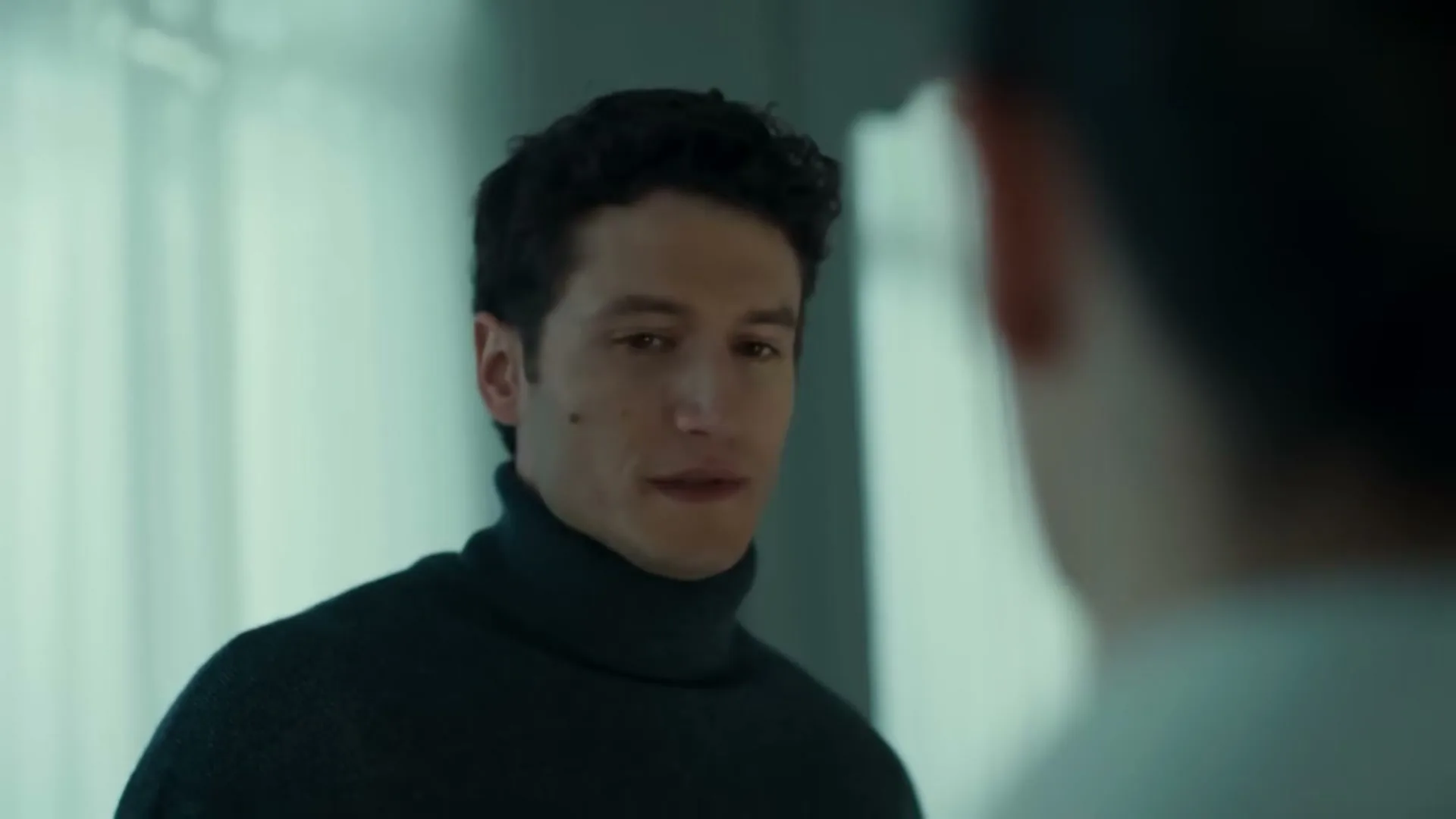 A young man with dark curly hair, wearing a dark turtleneck, looks at another person in a modern interior.