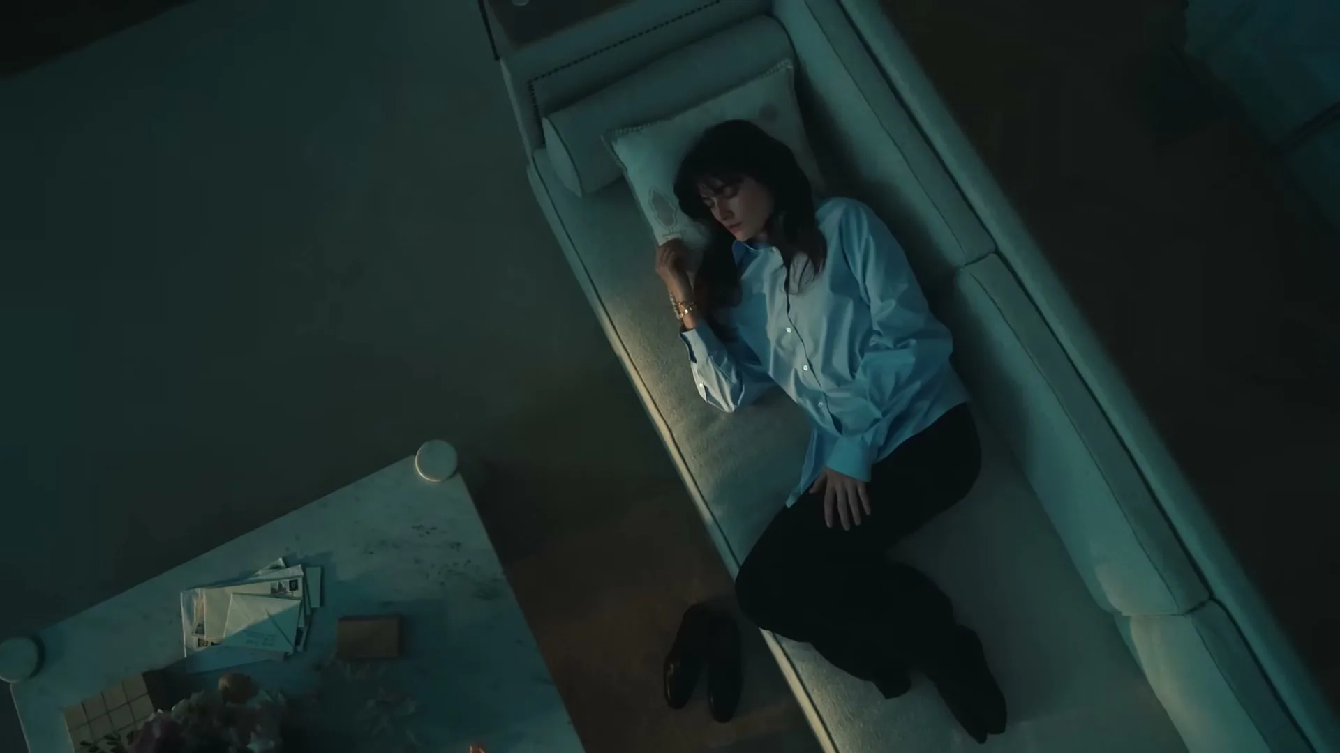 A woman with dark hair and a light blue shirt sleeps on a white couch in a dimly lit room, with a coffee table nearby.