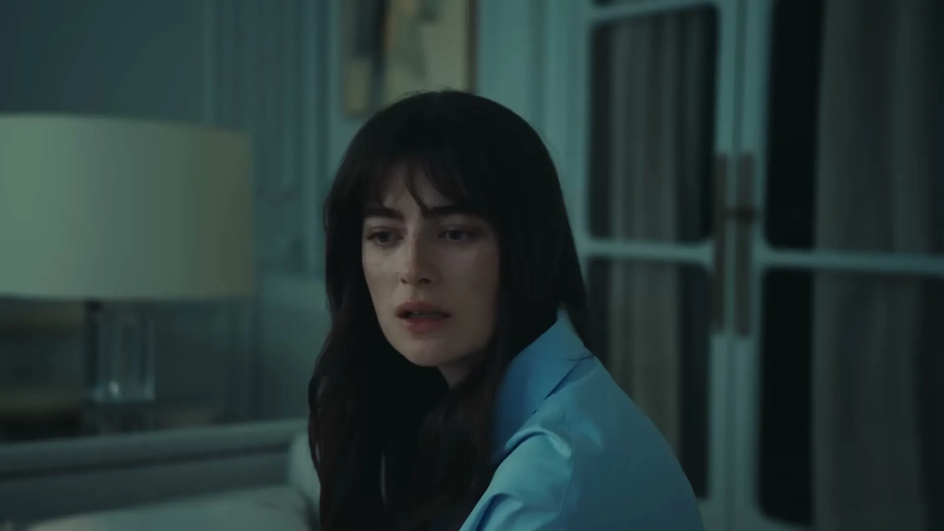 A young woman with dark hair and a blue shirt looks down with a concerned expression in a dimly lit room.
