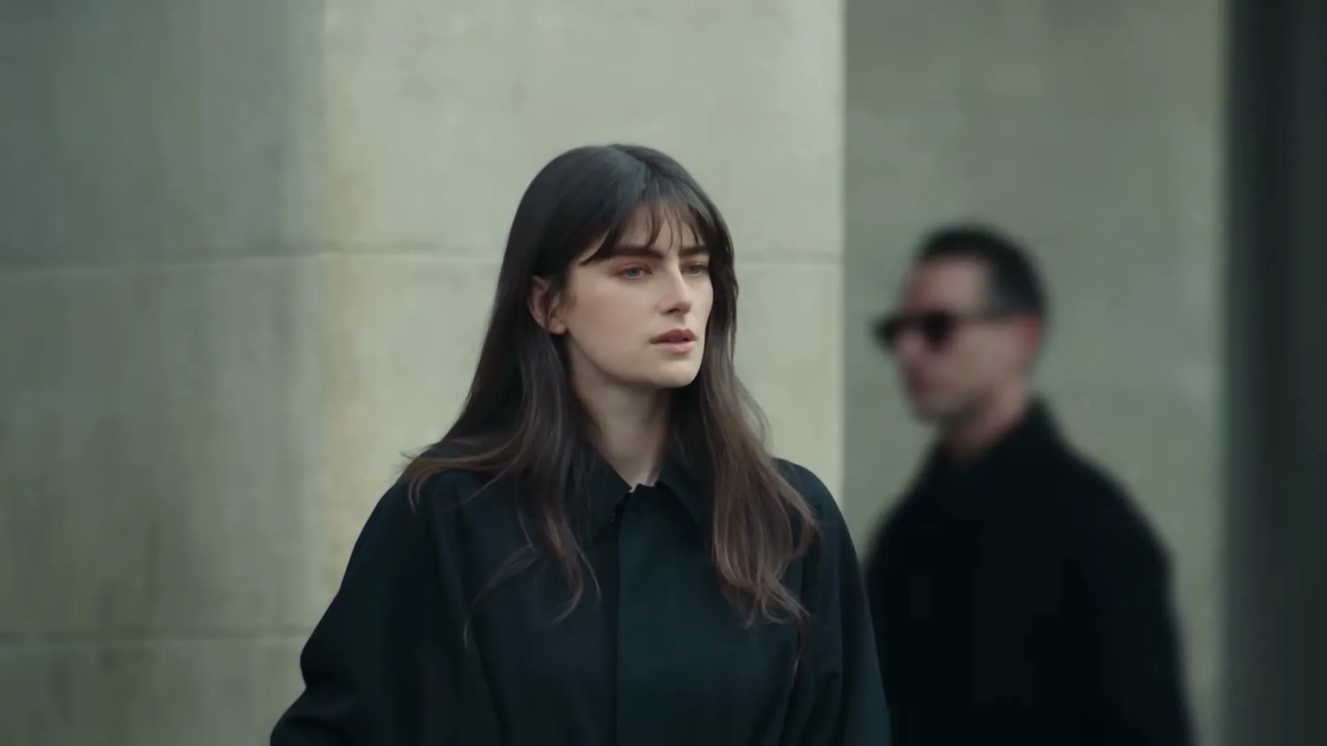 A young woman with dark hair and a black shirt looks off-camera, while a blurry man walks past in the background.
