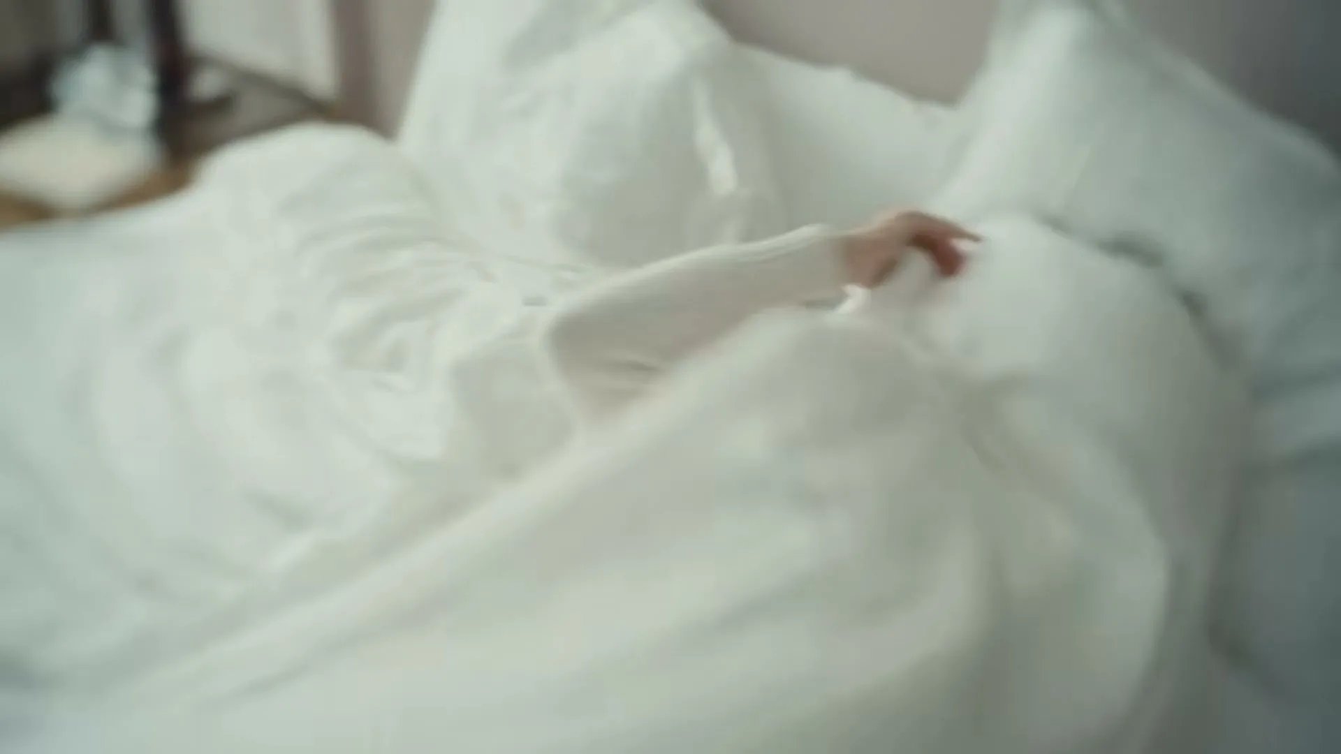 A young woman with dark hair wakes up in a white bed, laughing and smiling as she emerges from under the covers in a bright bedroom.