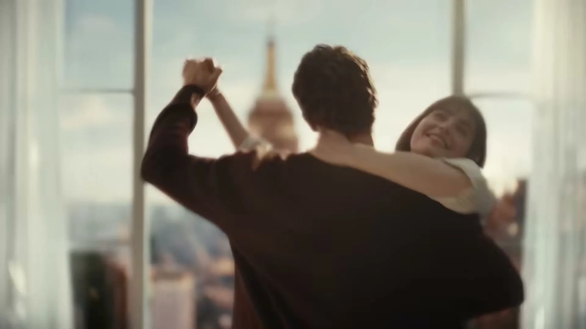 A young couple dances in a high-rise apartment with the Empire State Building visible through the window.