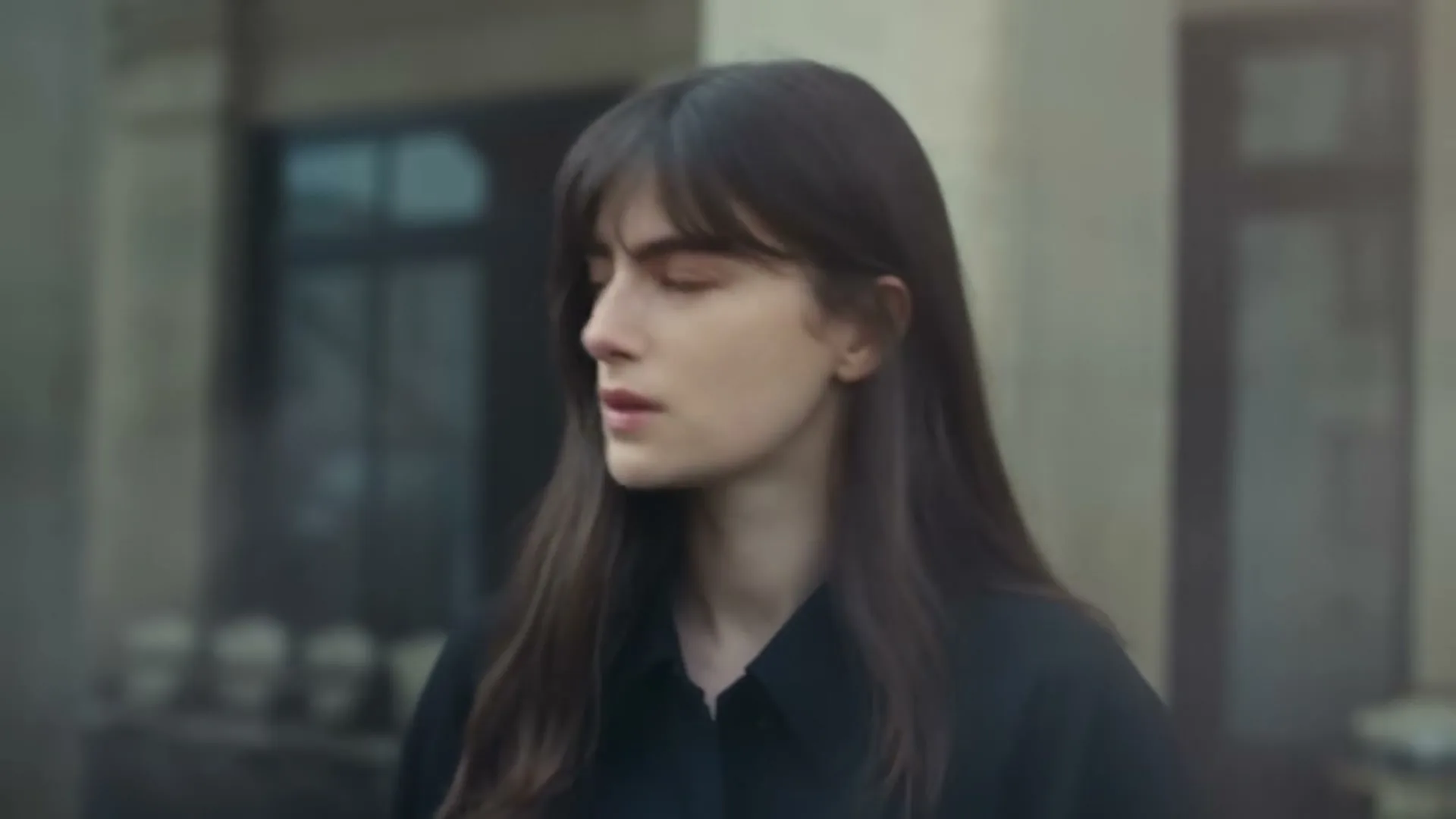 A young woman with long dark hair and bangs, wearing a black shirt, looks down and then turns her head to the right, outdoors.