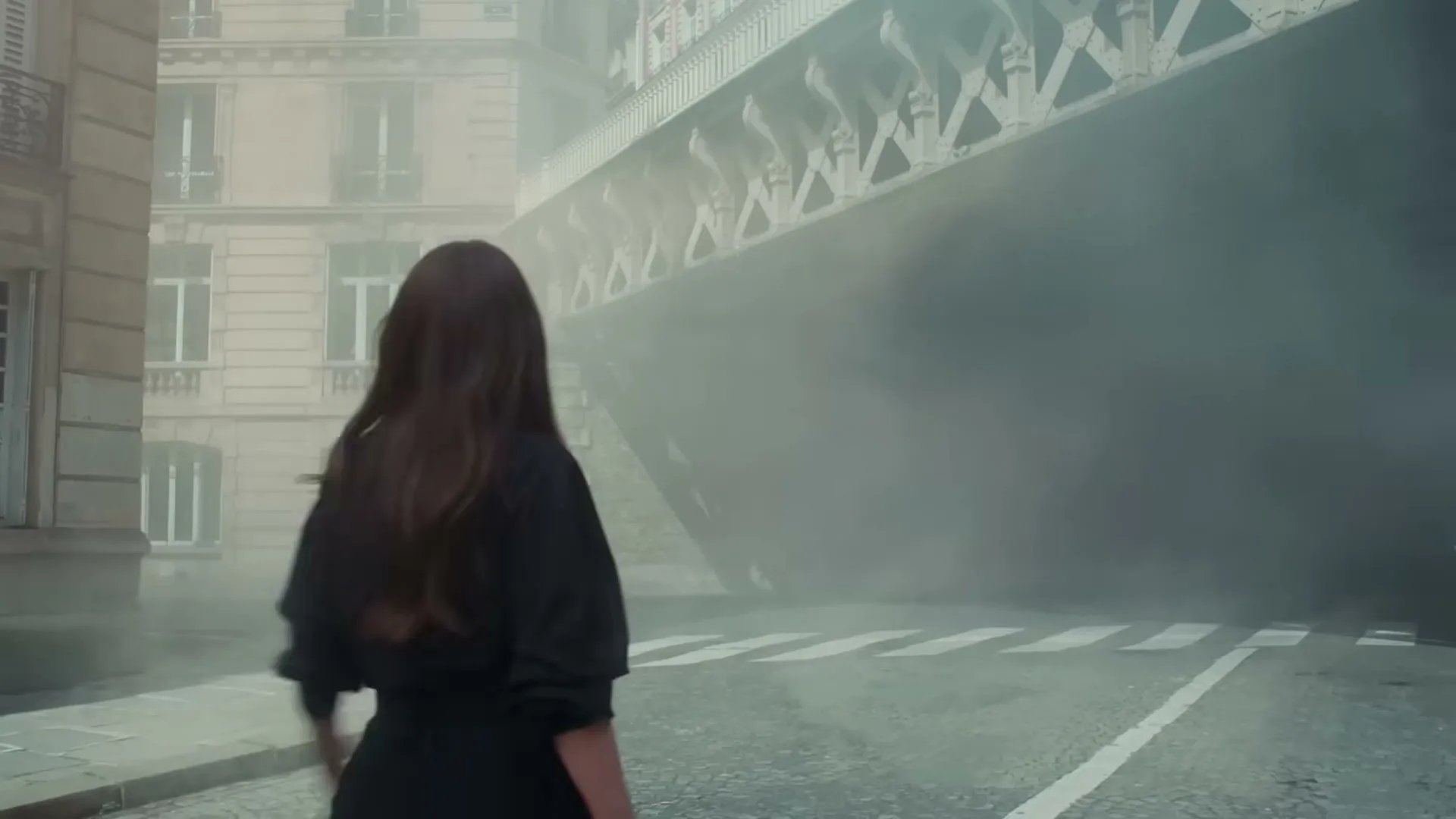 A woman with long dark hair, wearing a black dress, walks away from the camera on a misty street with a large bridge in the background.