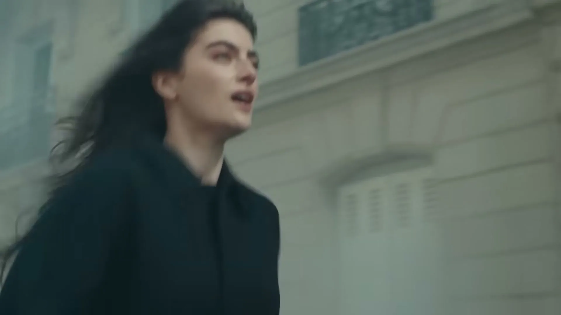 A young woman with dark hair and a black coat runs past a building, her hair blowing in the wind.