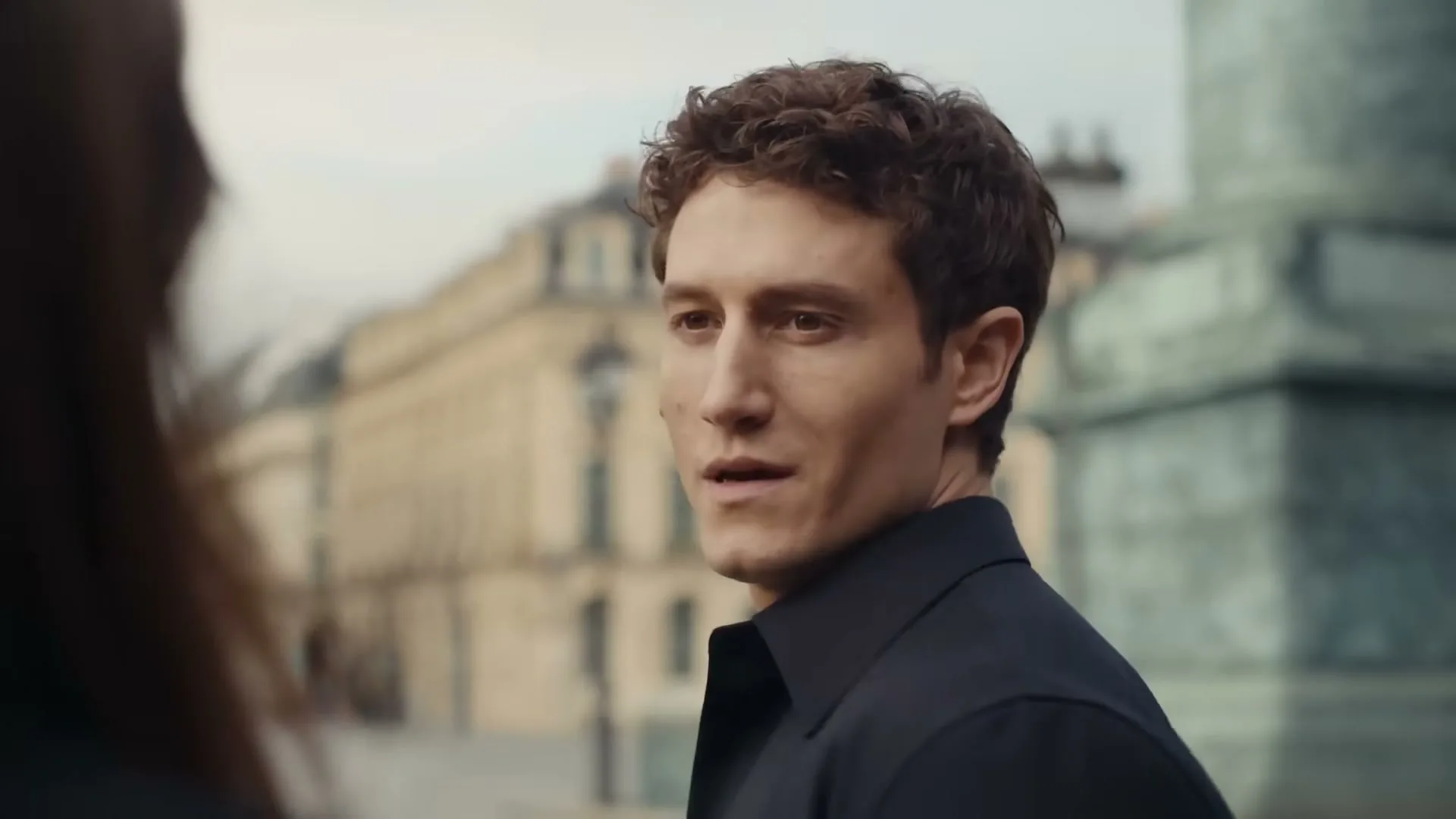 A man with curly brown hair in a dark shirt looks at a woman off-camera and smiles in a European city.