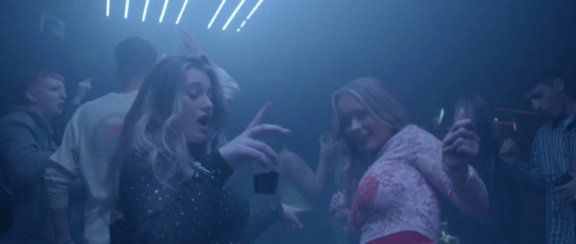 Two young women dance energetically in a dimly lit club, surrounded by other partygoers, with blue neon lights in the background.