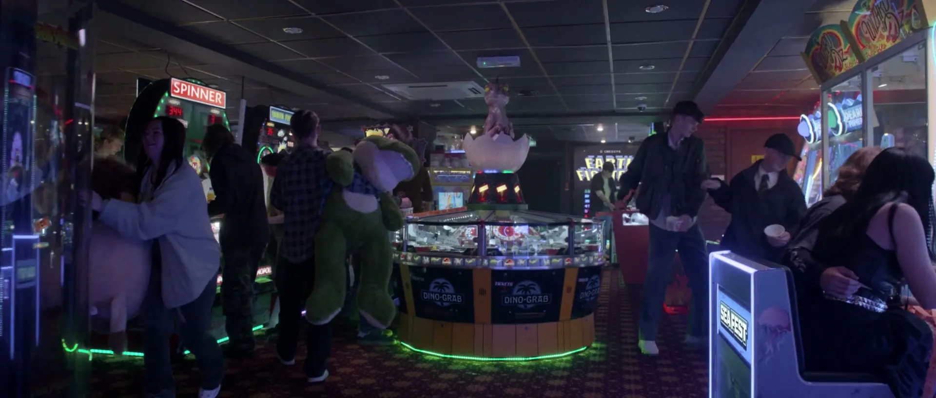 A group of young people are seen in an arcade, with one person carrying a large green dinosaur plush toy, as they walk around various game machines.