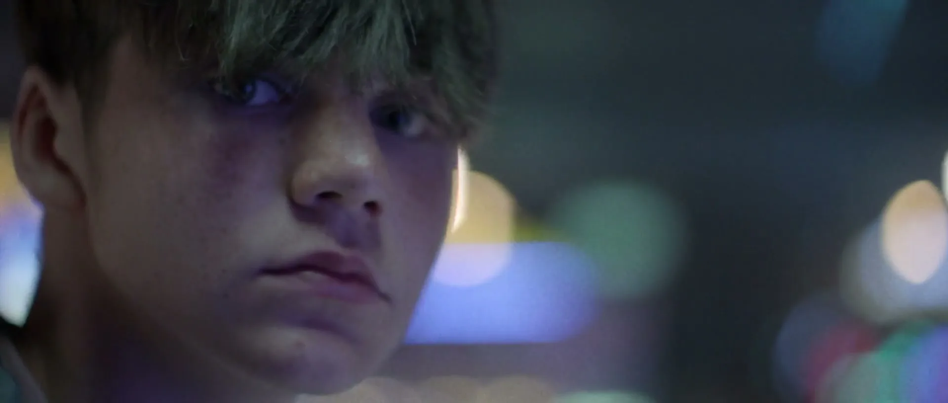 A young male with messy hair looks directly at the camera with a serious expression, illuminated by colorful lights in a dark setting.