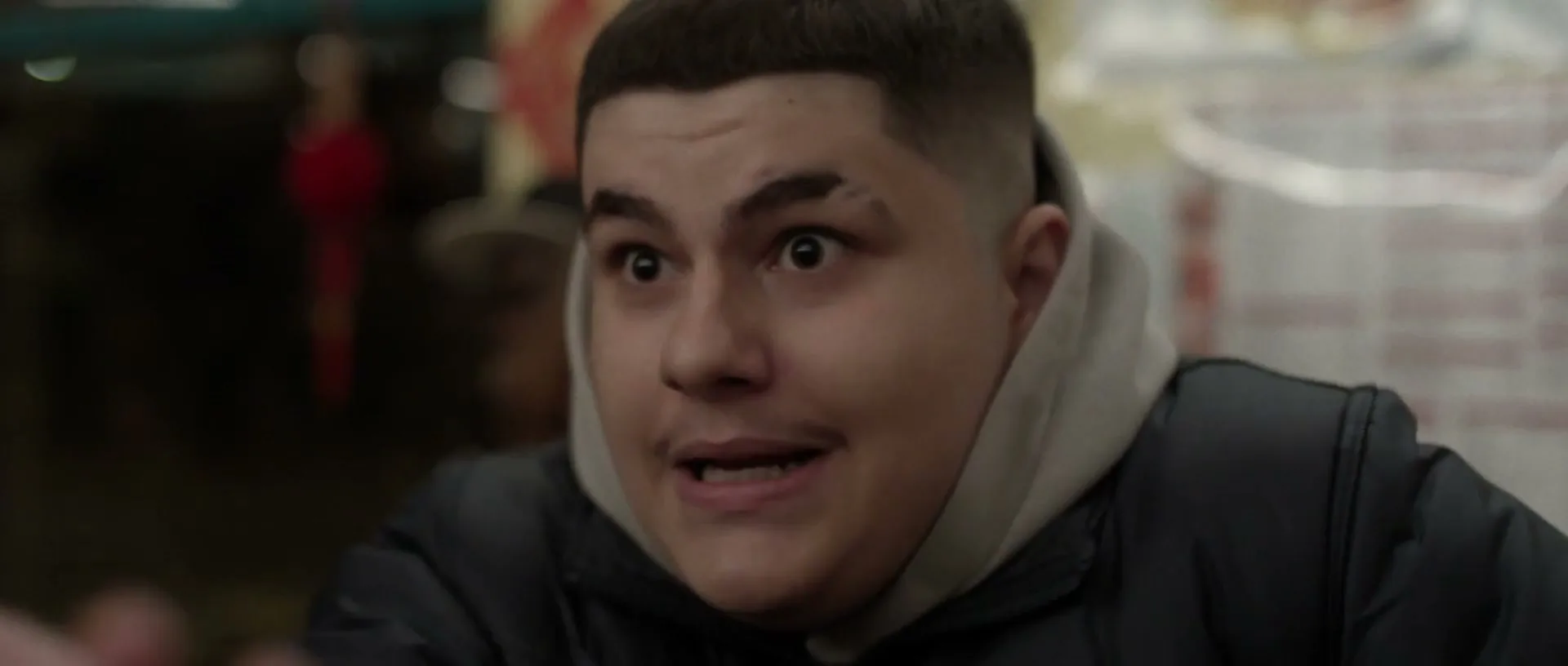 A young man with a fresh haircut and a light grey hoodie under a dark jacket looks surprised and speaks in a dimly lit outdoor setting.