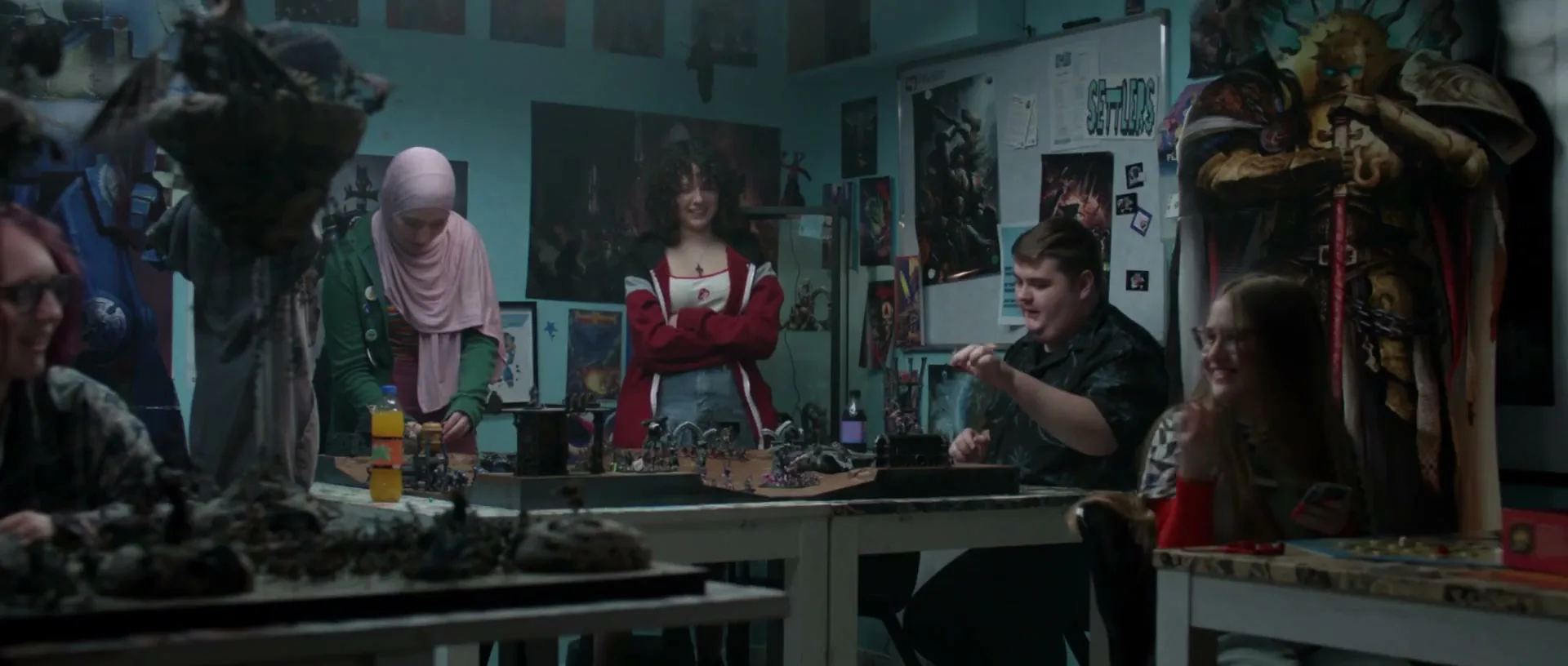 Four teens and young adults play a tabletop game with miniature figures in a dimly lit room filled with posters and game paraphernalia.