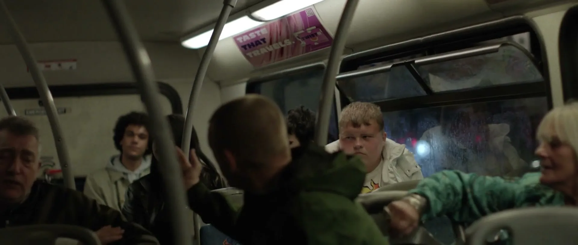 A diverse group of people, including a young man with blonde hair and a woman with white hair, are seated on a bus at night.