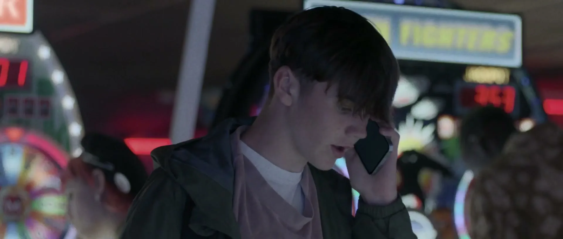 A young male teen with dark hair holds a phone to his ear, looking concerned in a dimly lit arcade.