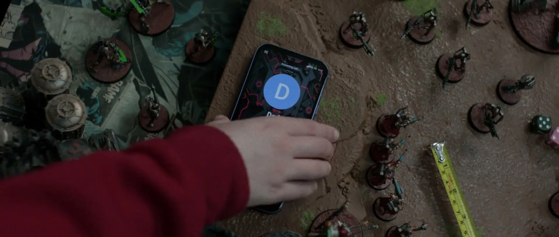 A child's hand in a red sleeve reaches for a smartphone displaying an incoming call from "Dad" amidst a tabletop wargame.