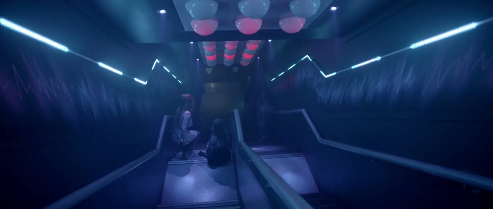 Two young women sit on stairs in a dark, neon-lit club entrance, while other people walk down the stairs behind them.