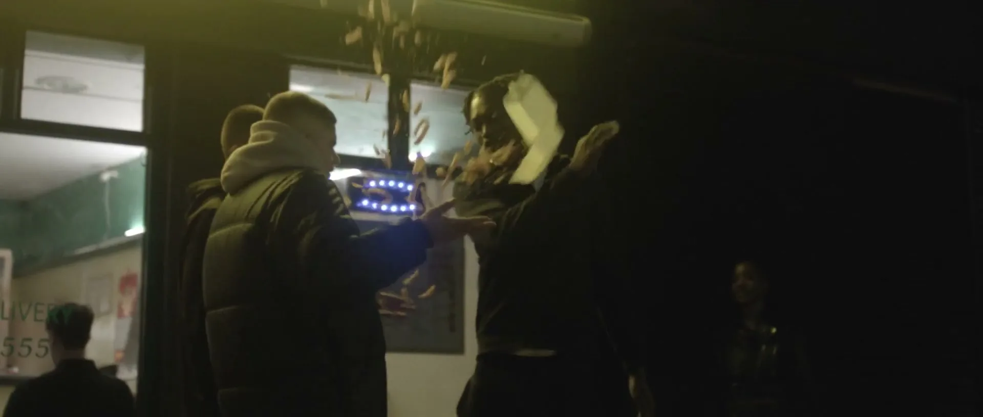 A young man in a dark hoodie throws a takeaway container of chips at another man outside a brightly lit takeaway shop at night.