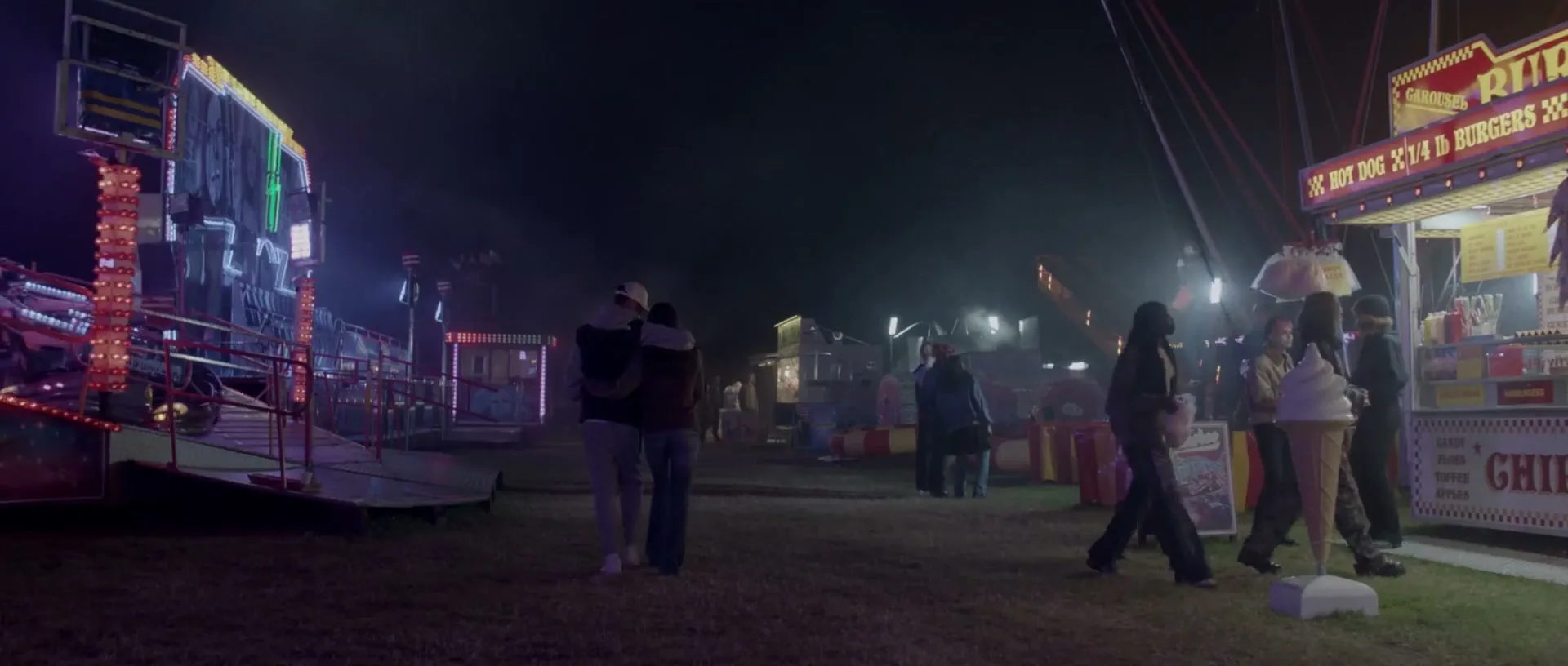 A young couple walks arm-in-arm through a dimly lit fairground at night, with rides and food stalls visible in the background.