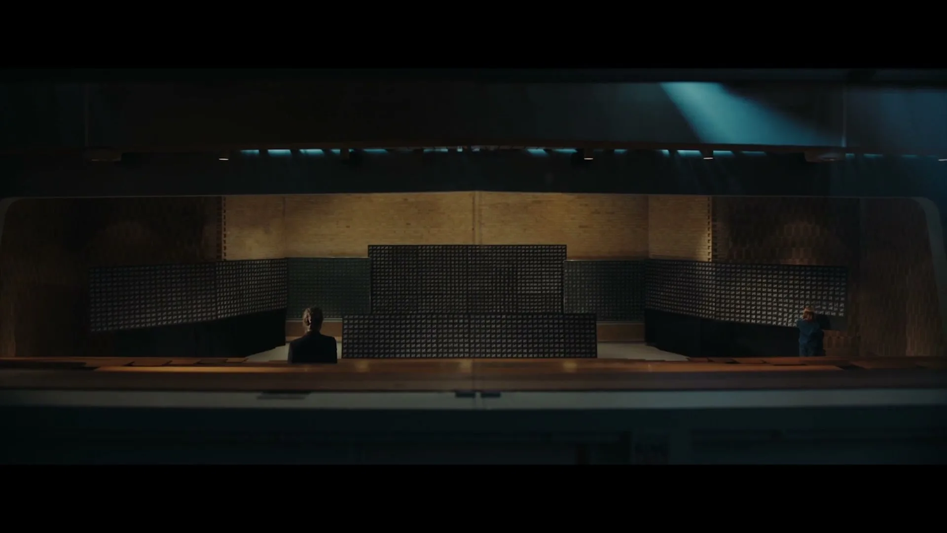 A man in a dark suit sits at a desk in a large, dark, soundproofed room, looking forward with a neutral expression.