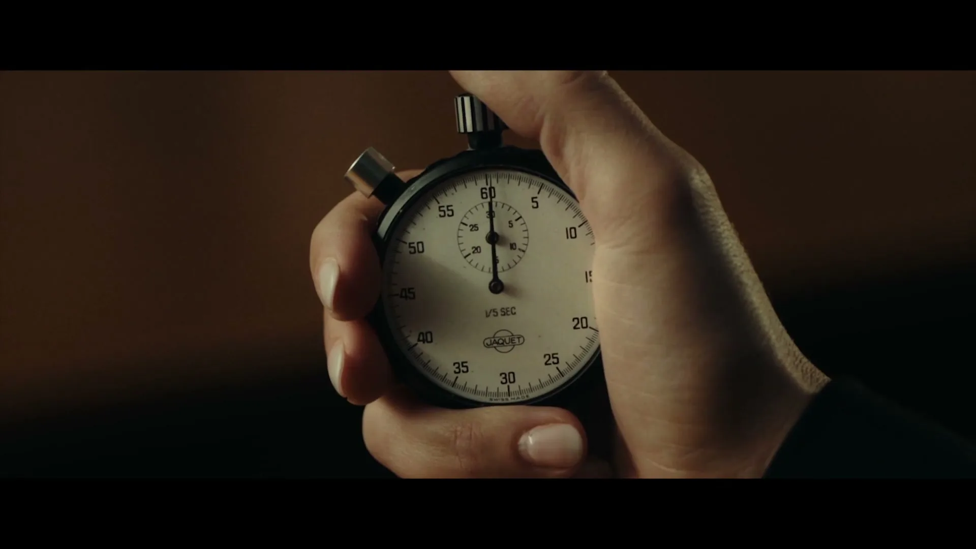 A female hand with manicured nails holds a stopwatch, pressing the button to start the timer, in a dimly lit interior.