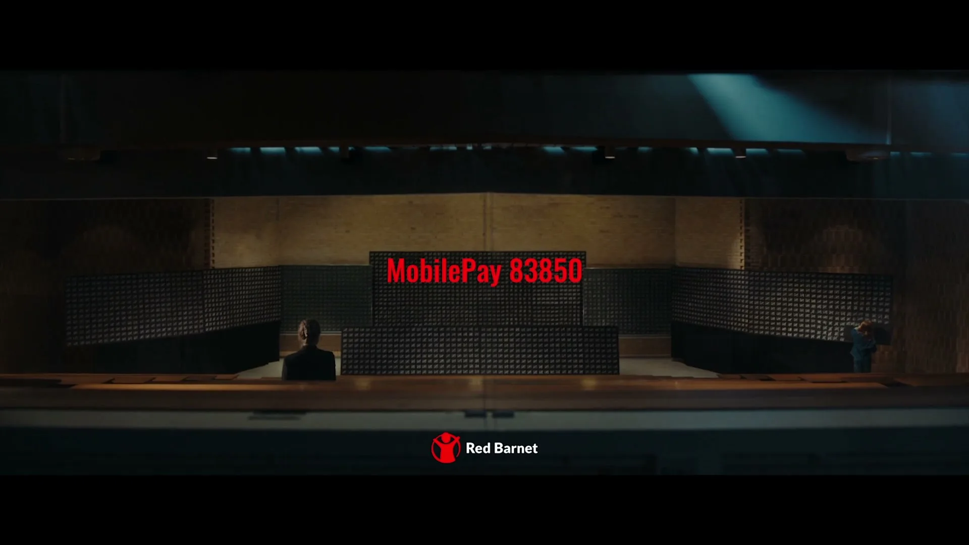 A wide shot of a dark, empty auditorium with two people sitting far apart, and red text and a logo appearing on screen.