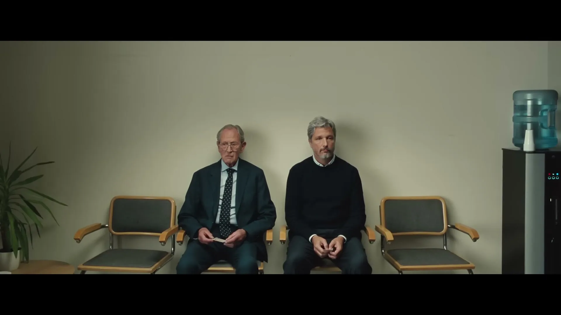 Two men, one older and one middle-aged, sit side-by-side in a waiting room, looking forward with neutral expressions.