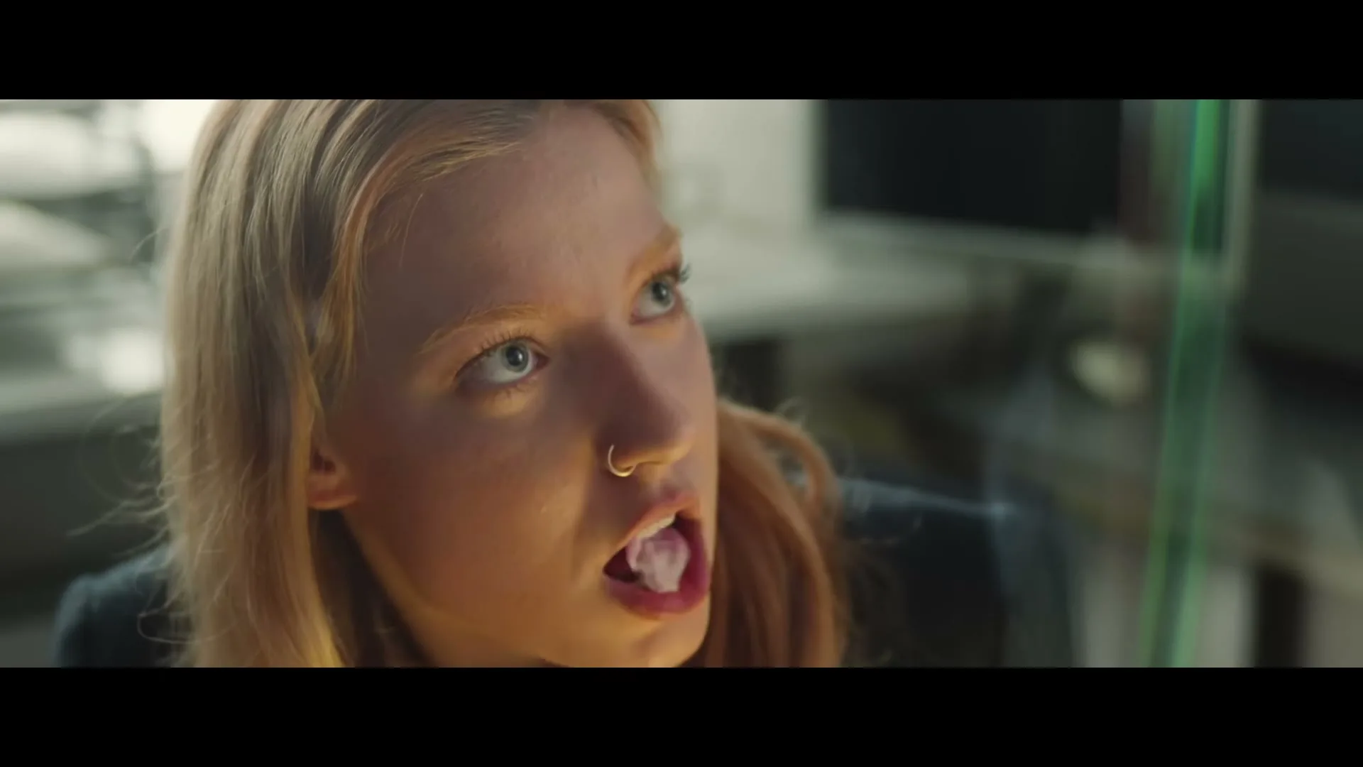 A young woman with blonde hair and a nose ring blows a bubble with gum, then it pops, in an office.