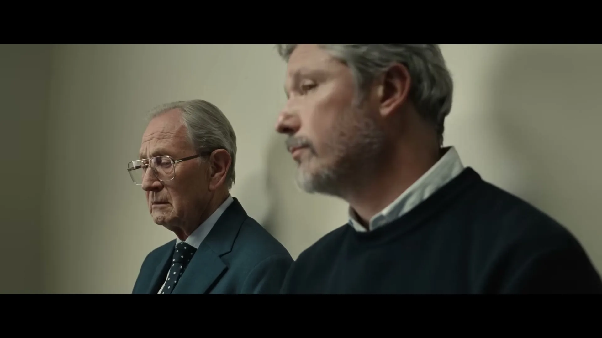 A middle-aged man with gray hair and beard looks forward, while an older man in a suit and glasses looks at him, in an interior setting.