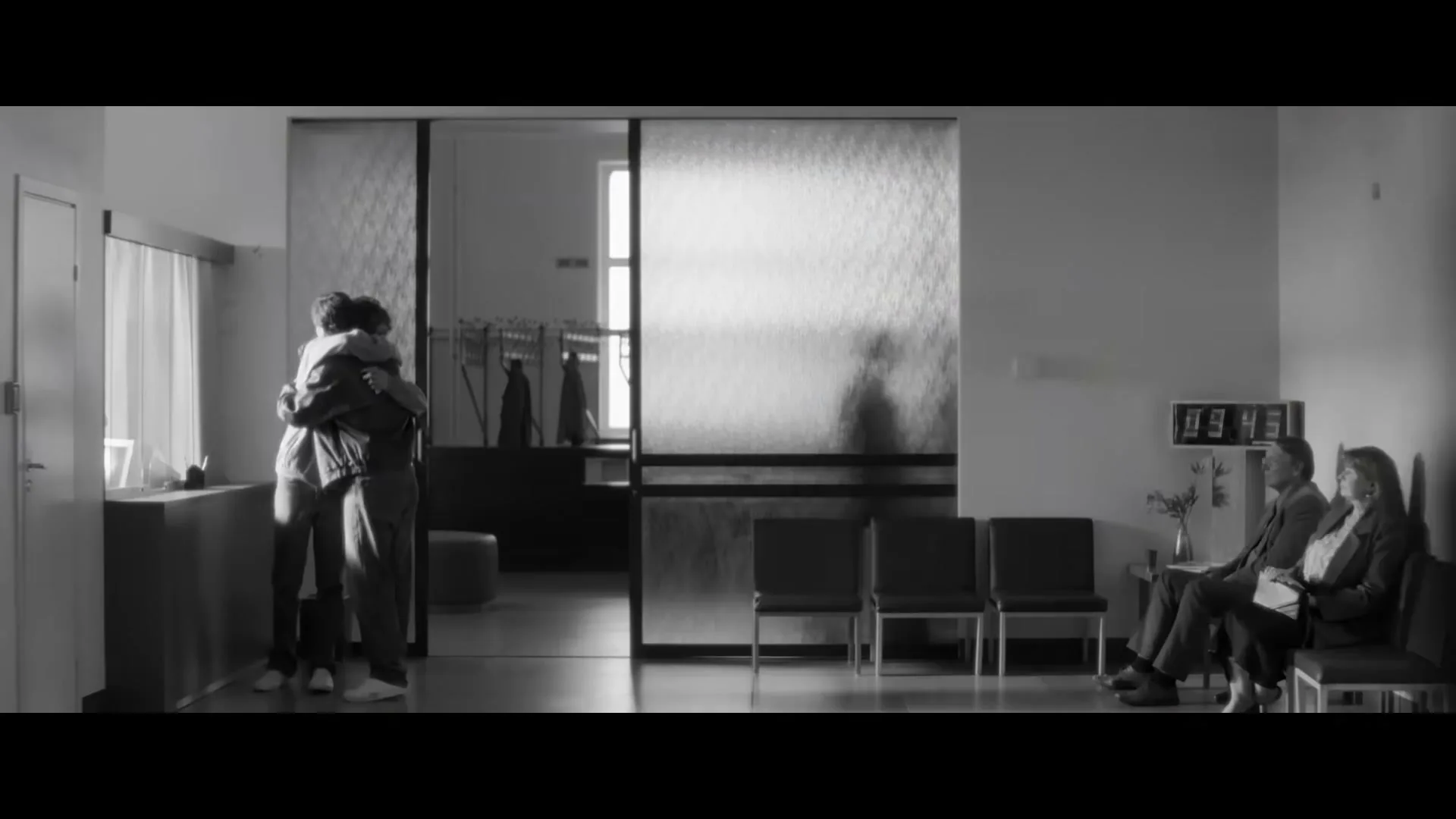 A father and son embrace in a hospital waiting room, while two other people sit nearby, in a black and white shot.