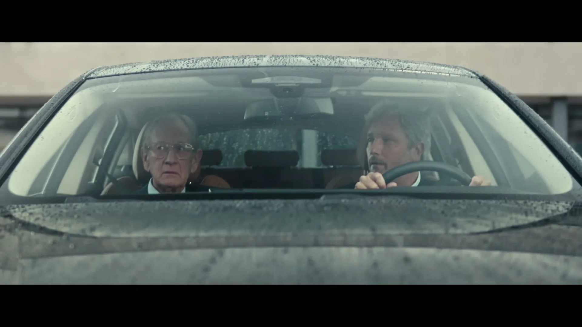 Two men, one older with glasses and one younger with a mustache, sit in a car, looking forward with serious expressions as rain falls on the windshield.