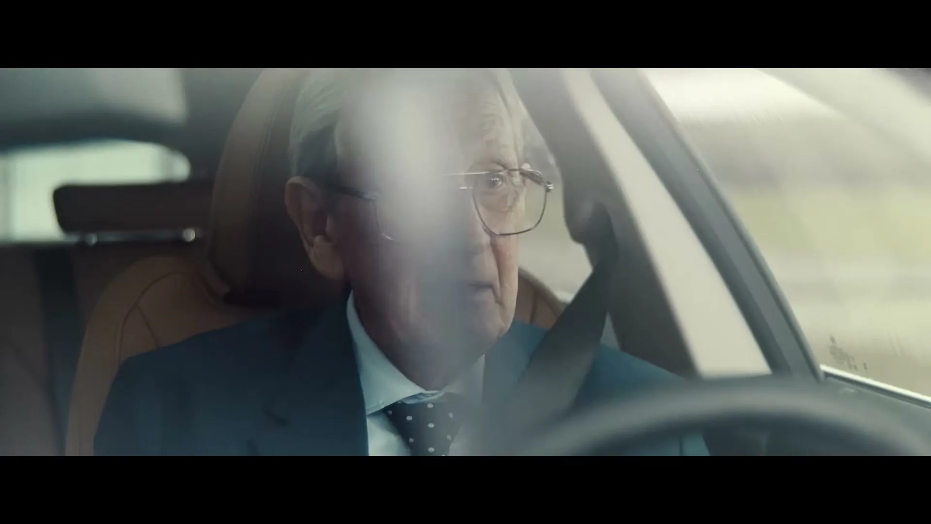 An elderly man in a suit and glasses drives a car, looking forward with a slightly surprised expression.