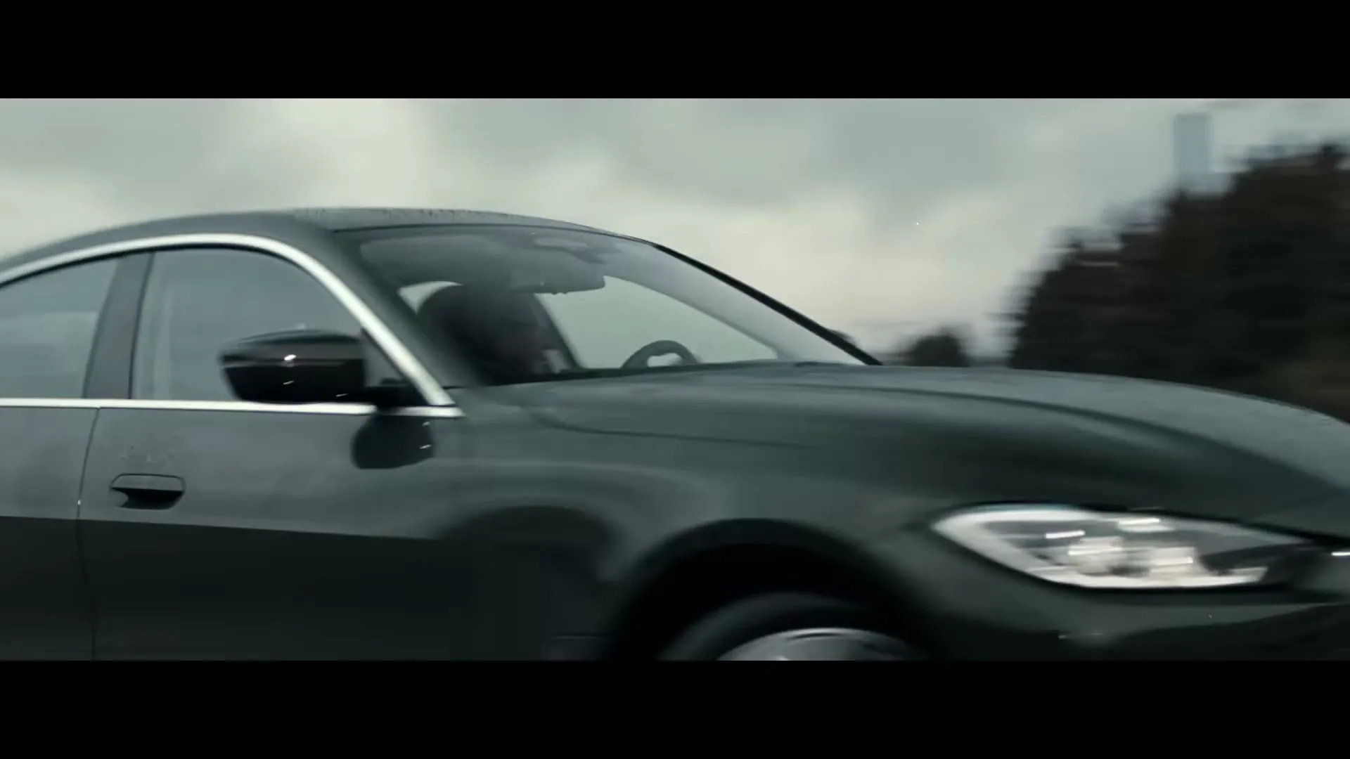 A dark green BMW car with two men inside drives on a road with buildings and trees in the background under an overcast sky.