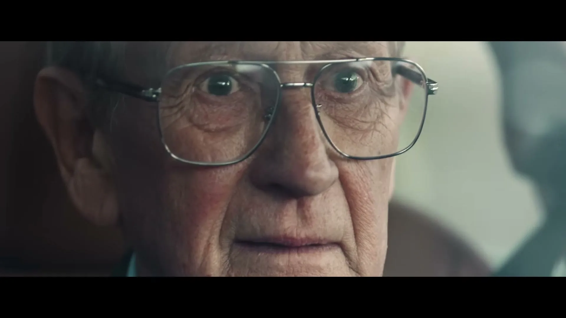 An elderly man with glasses looks surprised and then smiles, sitting in the passenger seat of a car.