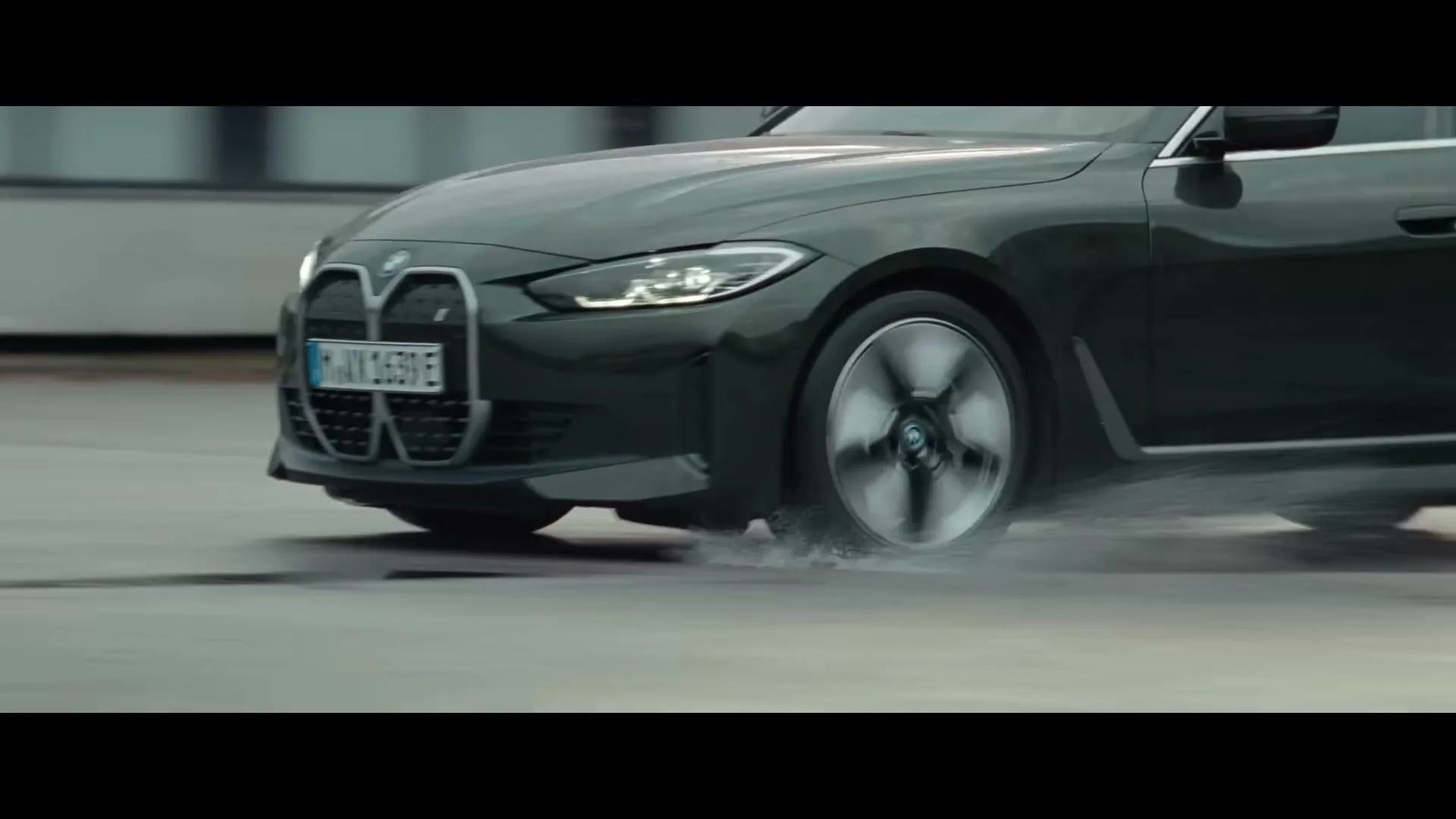 A dark green BMW car drives on a wet road, with water splashing from its tires, in an urban setting during the day.