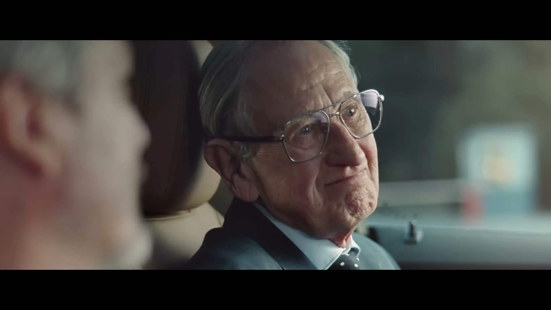 An elderly man with glasses and a suit smiles and looks at someone off-screen while sitting in a car during the day.