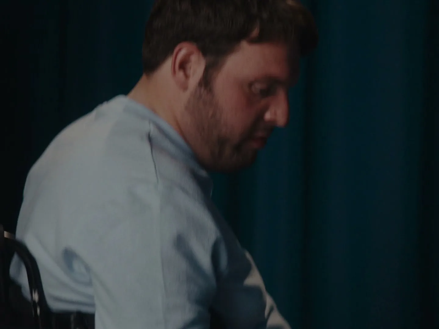 A man with a beard and light blue shirt looks around, then down, as another person walks behind him in a room with a blue curtain.