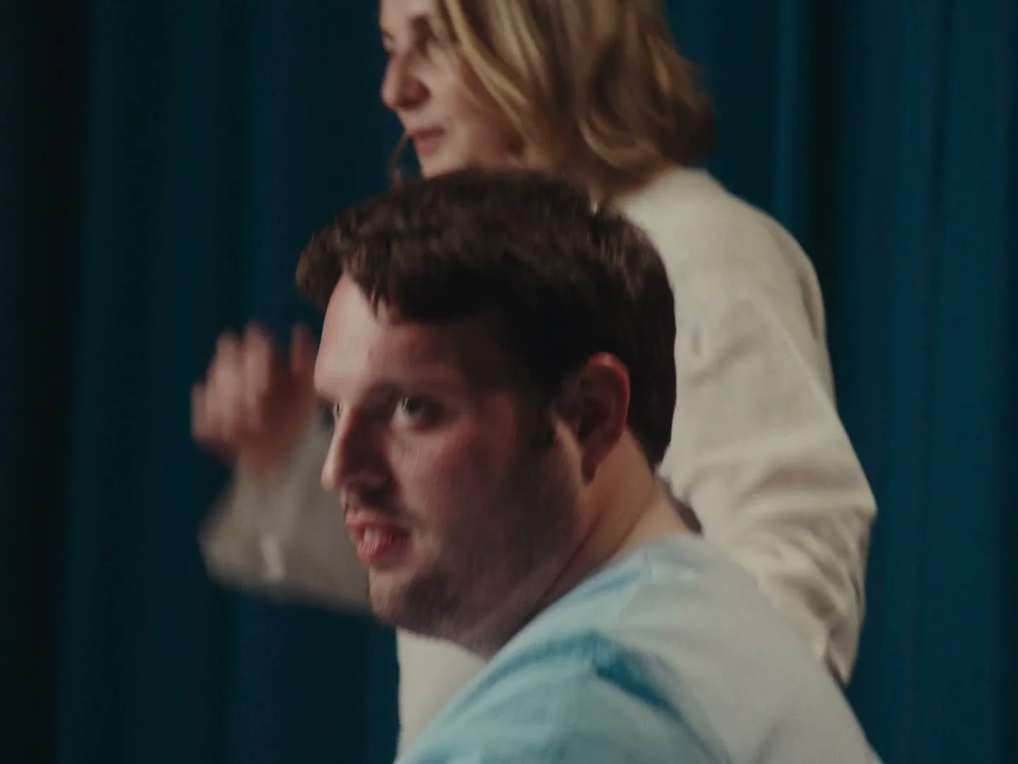 A man in a wheelchair looks at the camera as a woman walks past him in front of a blue curtain.