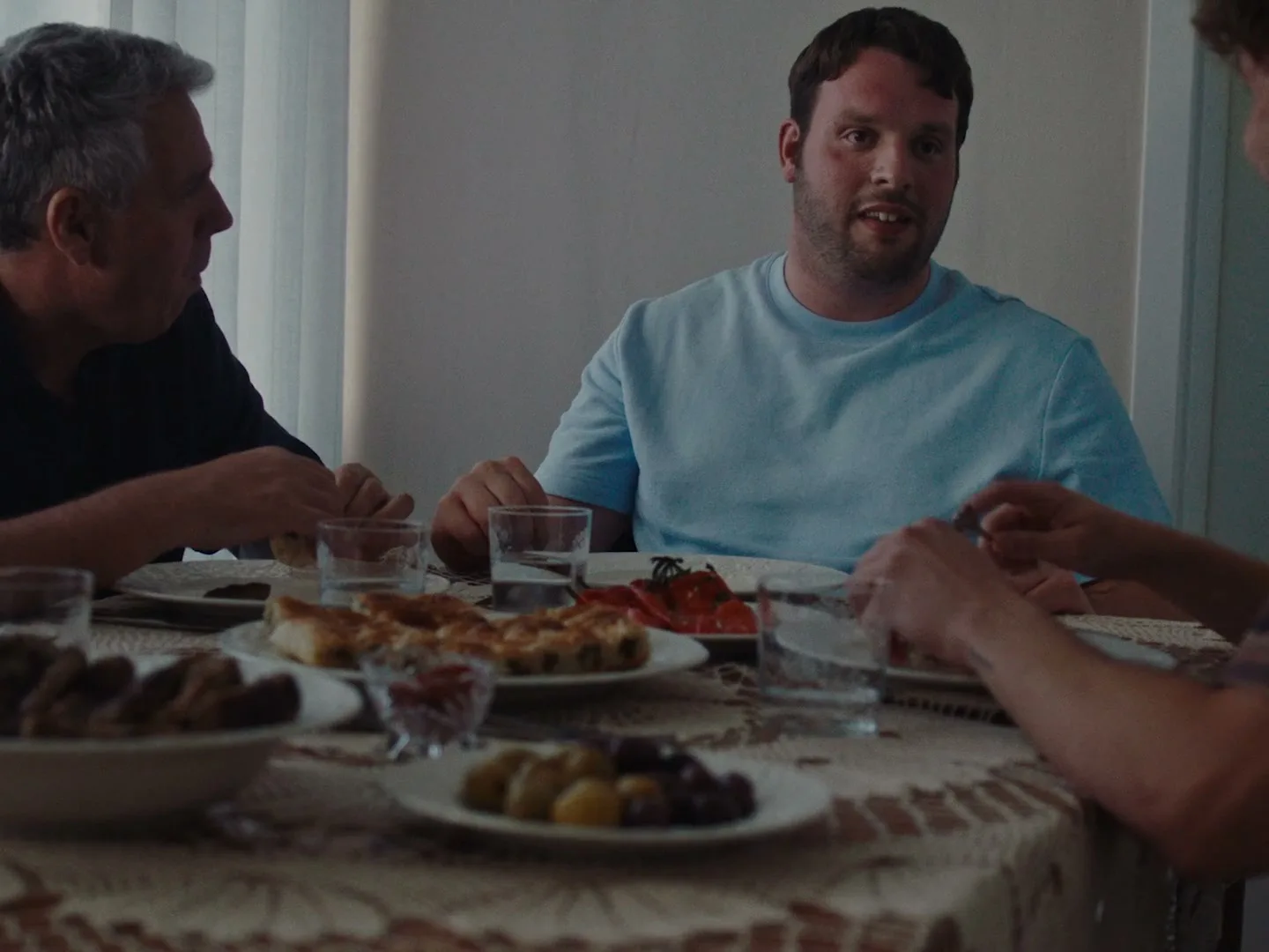A man in his 20s with a beard and light blue t-shirt talks animatedly at a dinner table with two other men, plates of food in front of them.