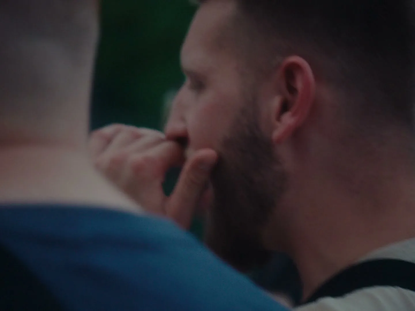 A man with a beard is seen in profile, biting his fingernails, with another person's shoulder in the foreground, outdoors.