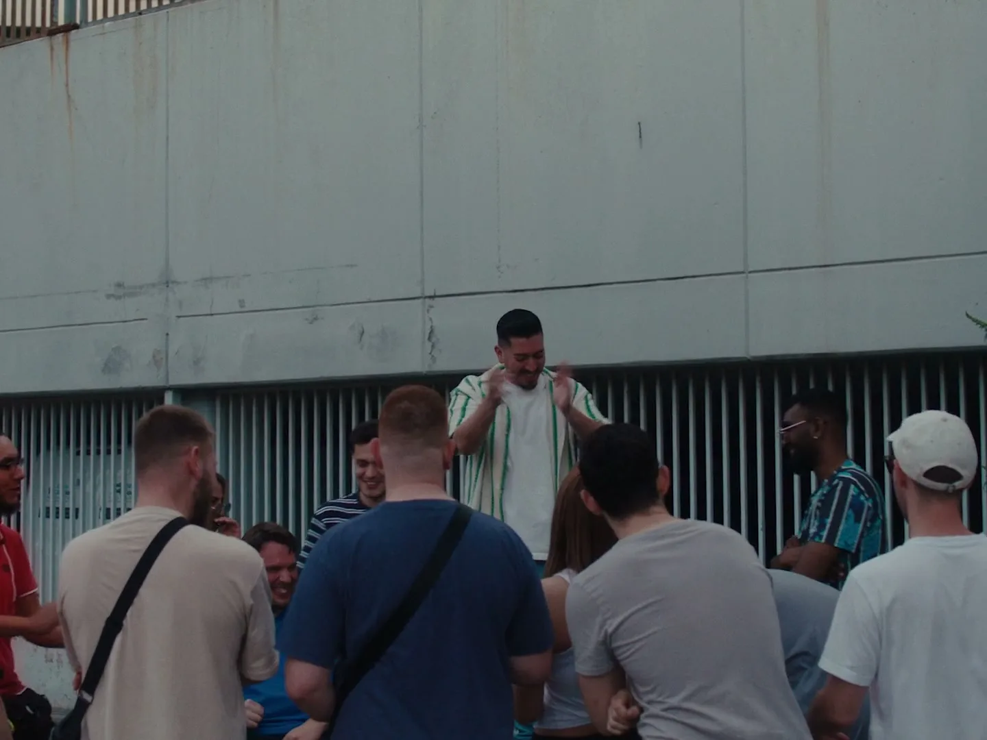 A diverse group of young adults gathers outdoors, watching a man in a striped shirt clap and gesture excitedly against a concrete wall.