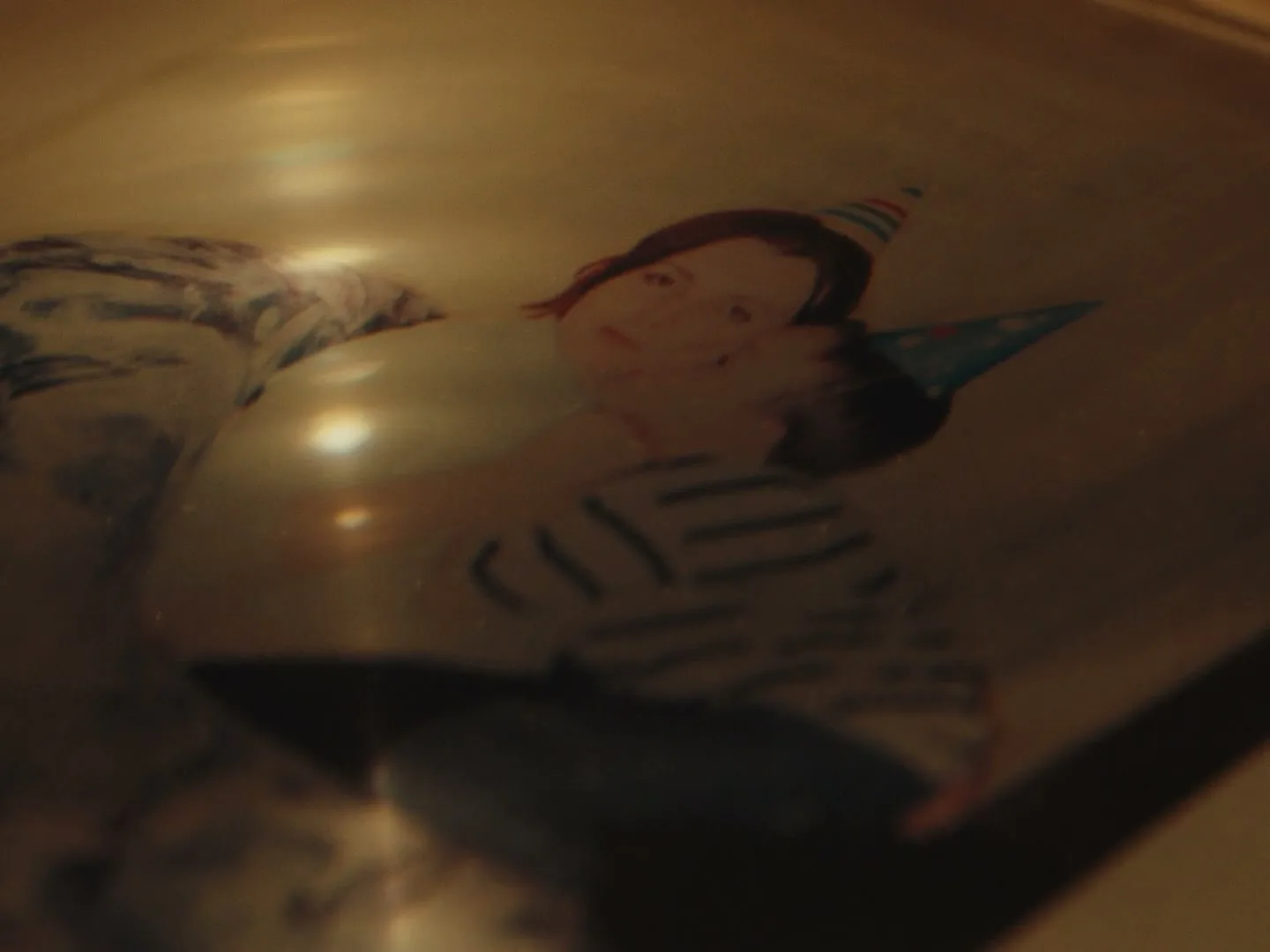 A blurry, distorted reflection of a woman holding a child, both wearing party hats, is seen on a shiny, metallic surface.