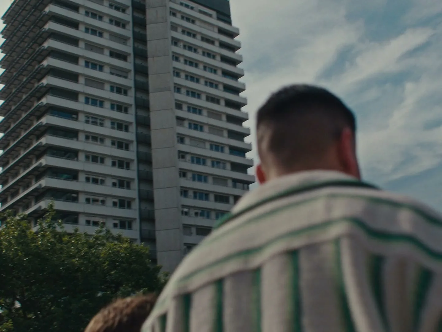 A man with a striped shirt, seen from behind, looks up at a tall, white apartment building under a cloudy sky.