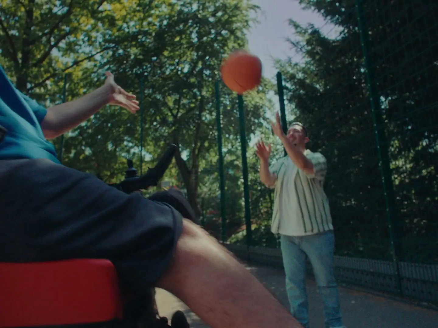 A man in a wheelchair plays basketball with another man on an outdoor court, catching a bright orange ball under a sunny sky.