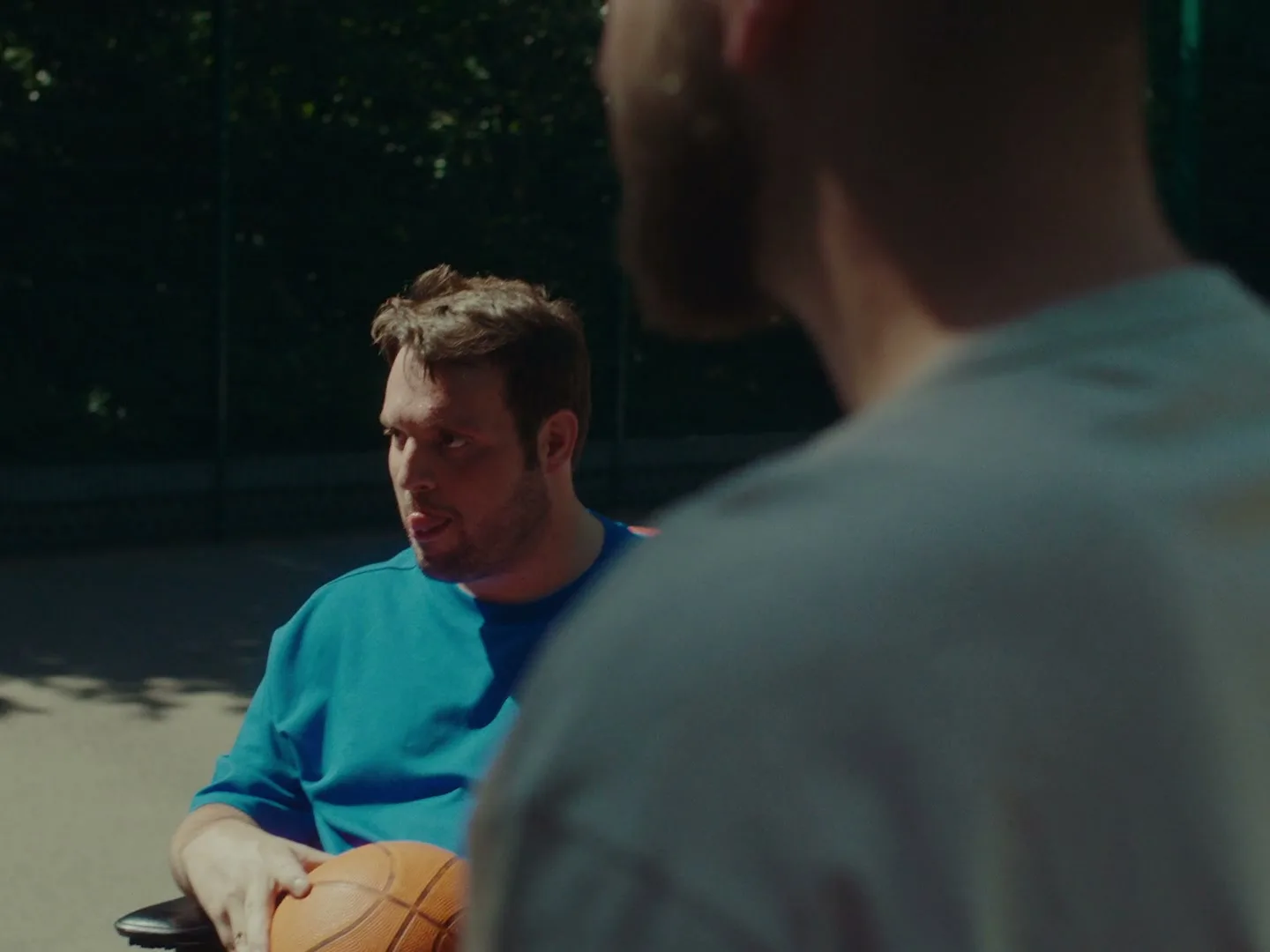 A man in a wheelchair, wearing a blue shirt, holds a basketball and talks to another man on an outdoor basketball court.